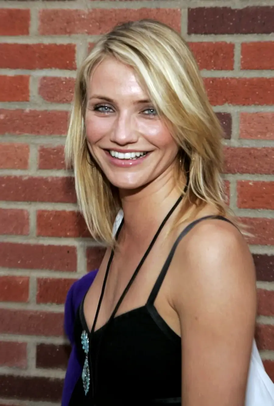 A woman with straight blonde hair smiles at the camera, standing in front of a red brick wall. She is wearing a black sleeveless top and a necklace.