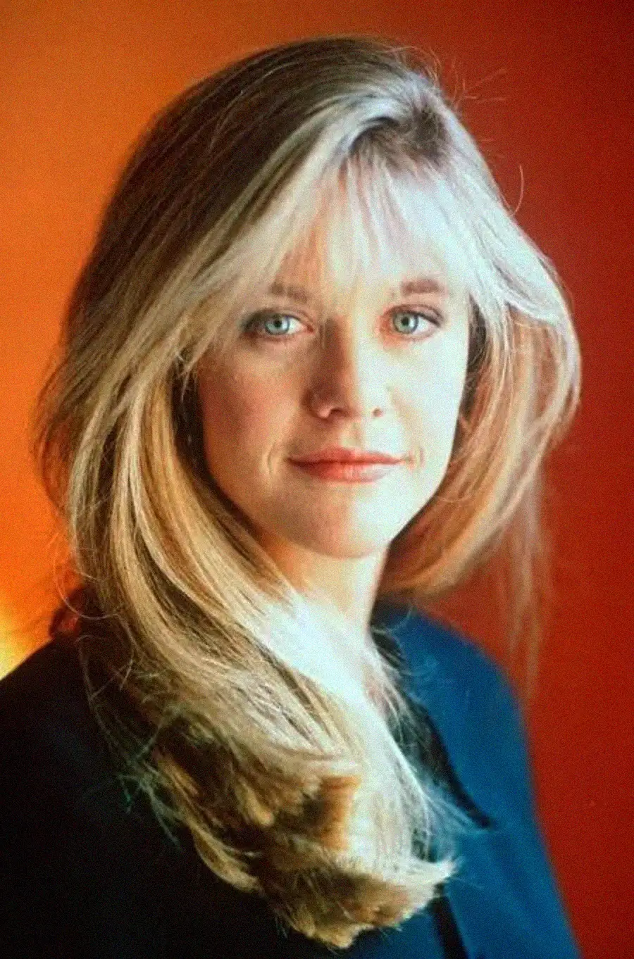 A woman with long blonde hair and blue eyes is wearing a dark top, posing against a warm orange background, looking confidently at the camera with a slight smile.