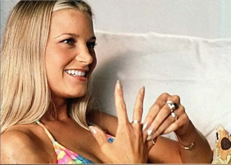A smiling woman with long blonde hair and a colorful swimsuit sits on a white couch, showing off rings on her fingers and looking to the side.