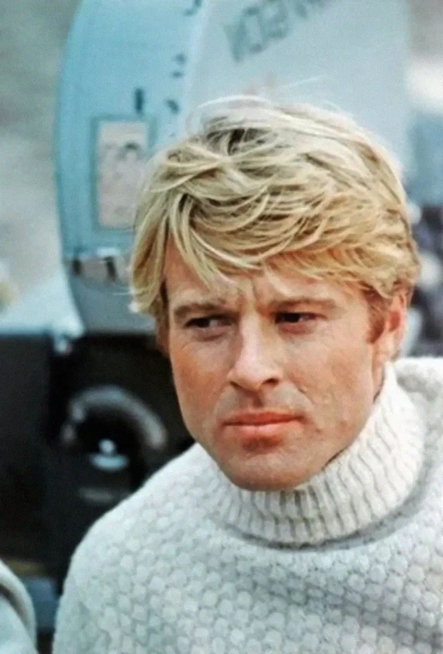 A man with blond hair wearing a white turtleneck sweater looks to the side, with a serious expression. A vintage film camera is visible in the background.