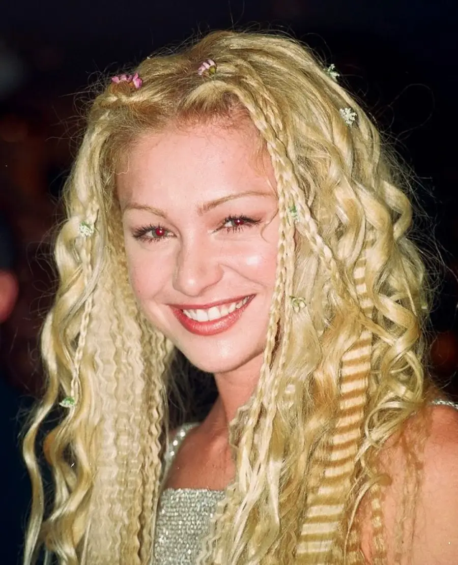 A smiling woman with long, blonde crimped and braided hair decorated with small colorful clips, wearing a sparkly light-colored top, posing against a dark background.