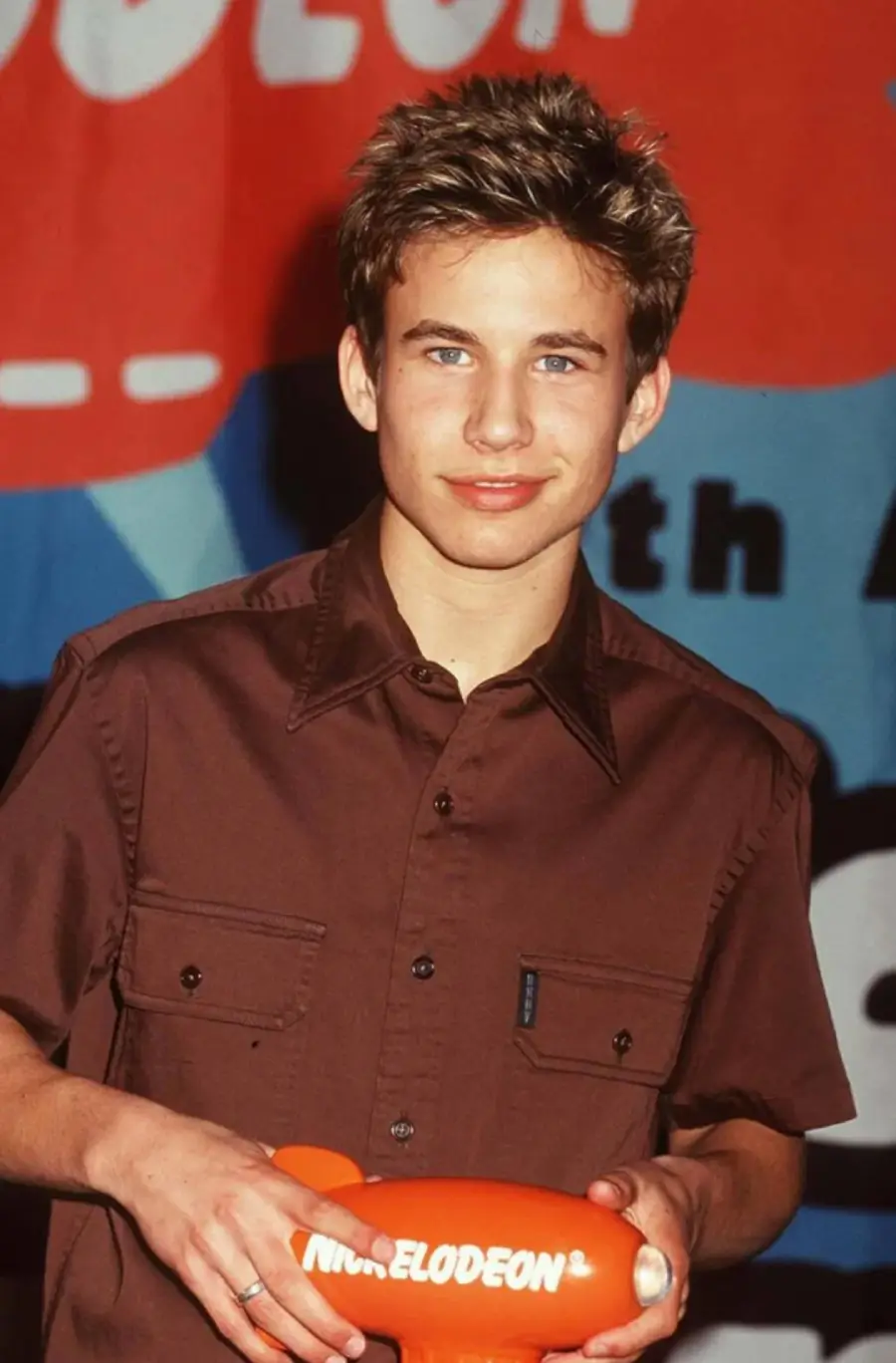 A young man with short, spiked hair holds an orange Nickelodeon award. He wears a brown button-up shirt and stands in front of a colorful Nickelodeon-themed backdrop.