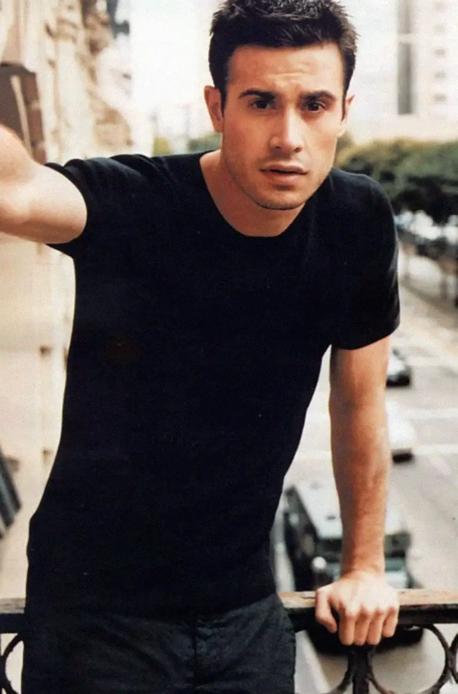 A man with short dark hair wearing a black t-shirt leans on a balcony railing, looking toward the camera. A city street with cars and trees is visible in the blurred background.