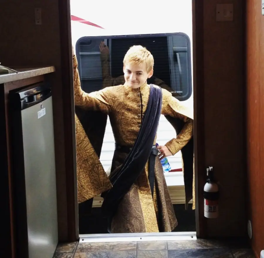 A person with short blond hair wearing an ornate gold and brown costume and a dark sash stands in a doorway, smiling and holding a water bottle, with a white vehicle visible outside behind them.