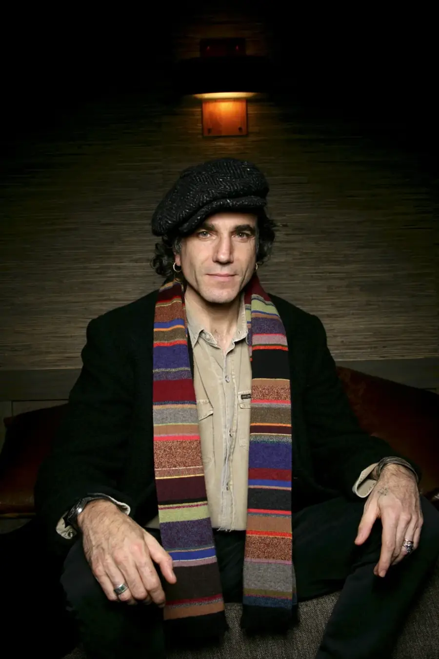 A man with curly hair wearing a flat cap, beige shirt, dark coat, and a colorful striped scarf sits with his hands resting on his knees against a dimly lit, textured background.