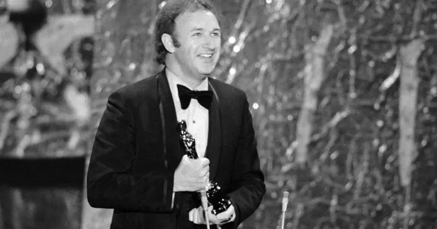 A man in a tuxedo smiles while holding an Oscar statuette on stage, with a textured backdrop behind him. The photo is in black and white.