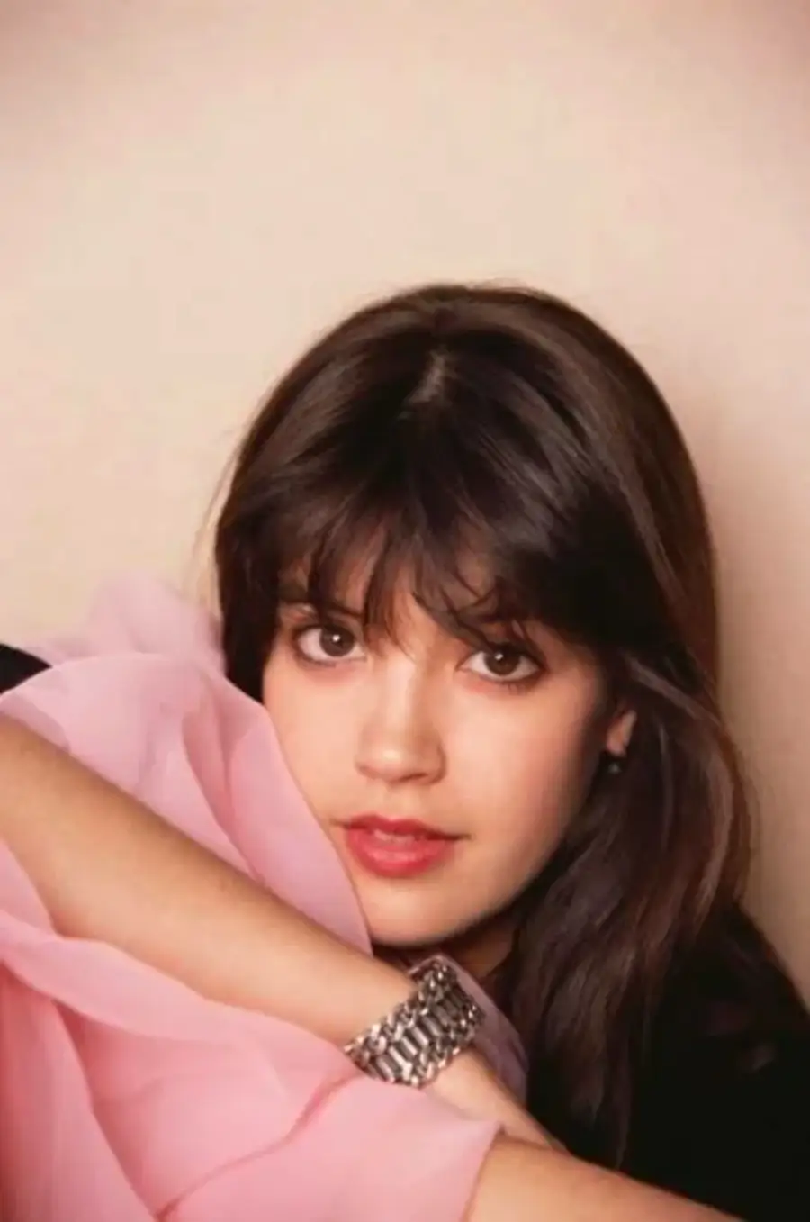 A young woman with long dark hair and bangs looks at the camera, resting her arm on a pink, sheer fabric. She is wearing a silver bracelet and has a soft, relaxed expression against a plain background.