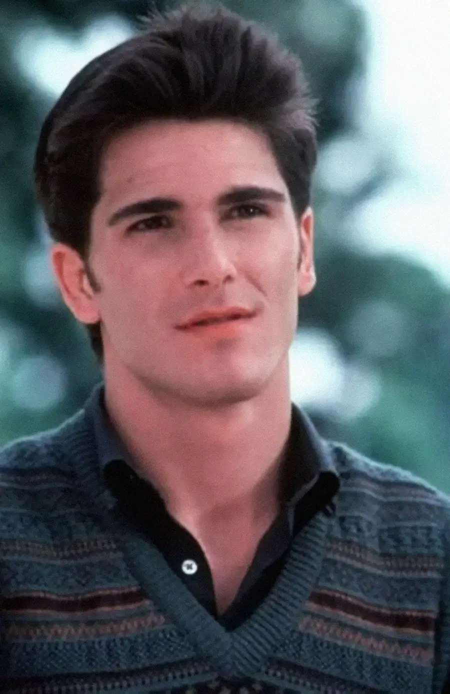 A young man with dark hair, wearing a patterned sweater over a collared shirt, looks slightly to the side with a neutral expression. The background is blurred greenery.