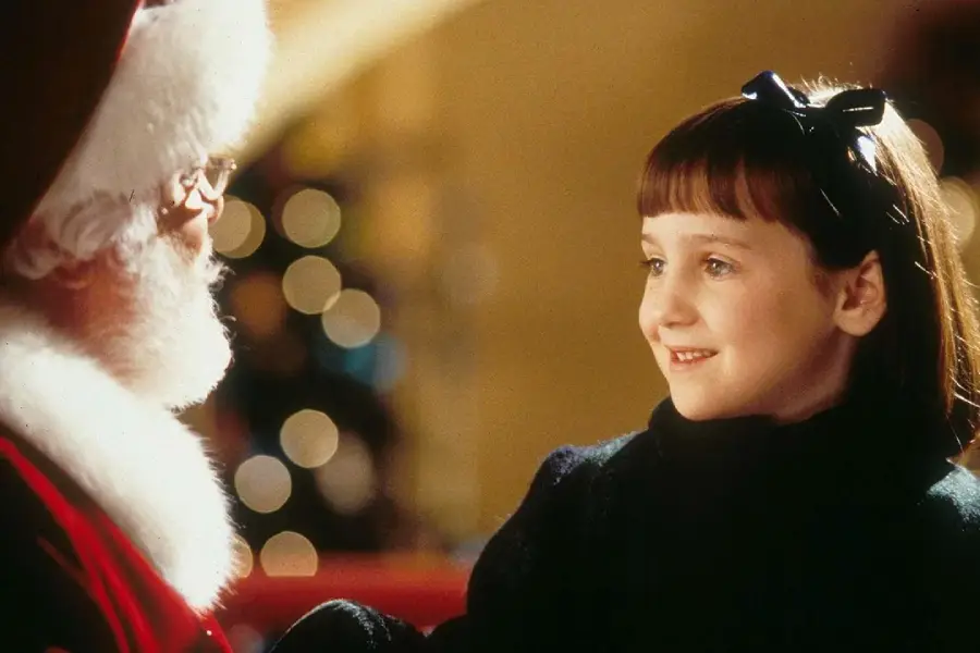 A young girl with brown hair and a black bow smiles warmly at a man dressed as Santa Claus, who has a white beard and glasses. Soft, festive lights glow in the blurred background.