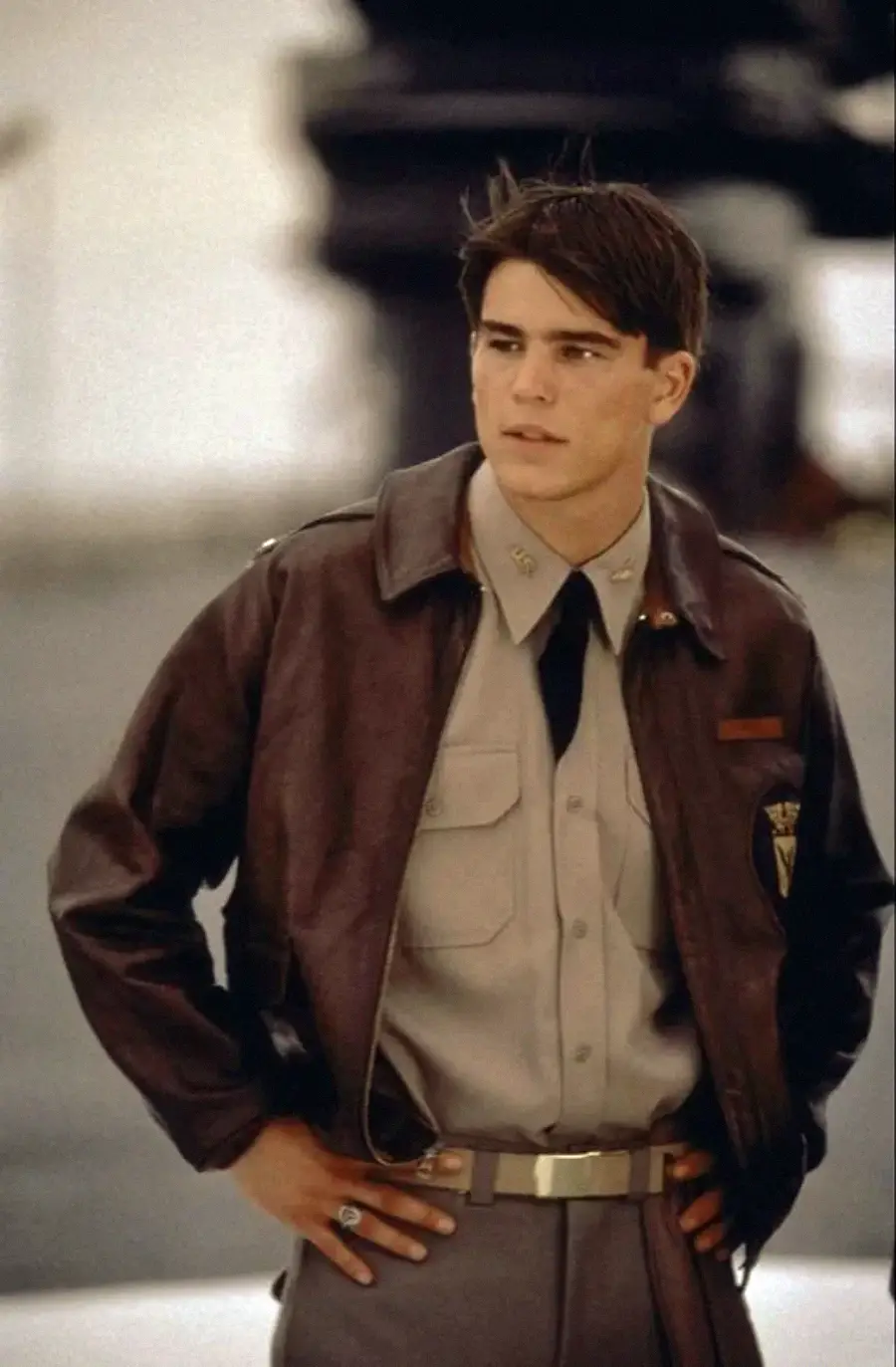 A young man in a military uniform with a leather bomber jacket stands outdoors with his hands on his hips, looking to the side. His short dark hair is neatly styled and he appears thoughtful.