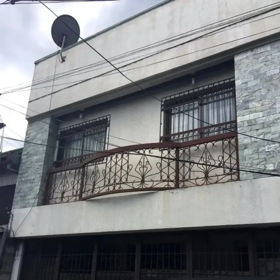 A two-story building with metal railings on a small balcony, bars on the windows, stone tiles on one side, and a satellite dish attached near the roof. Power lines are visible crossing in front.