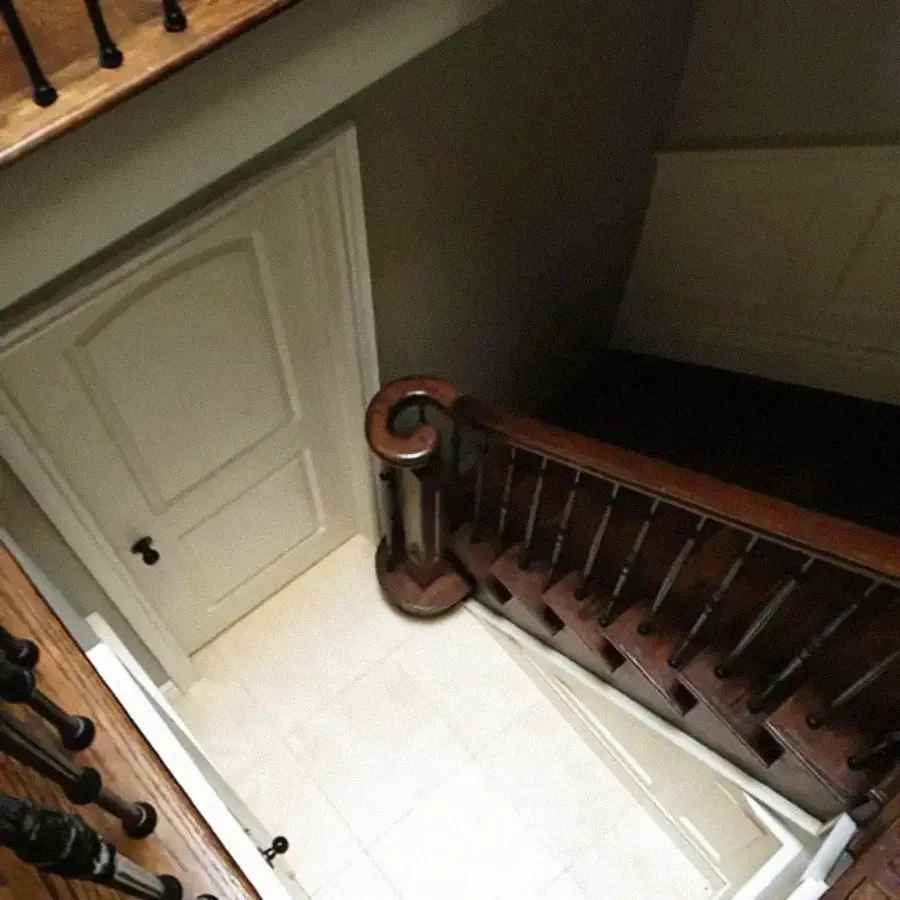 View from above of a staircase with dark wooden railing and white spindles, leading down to a landing with a closed white door and light-colored tile floor. Walls are painted a neutral shade.