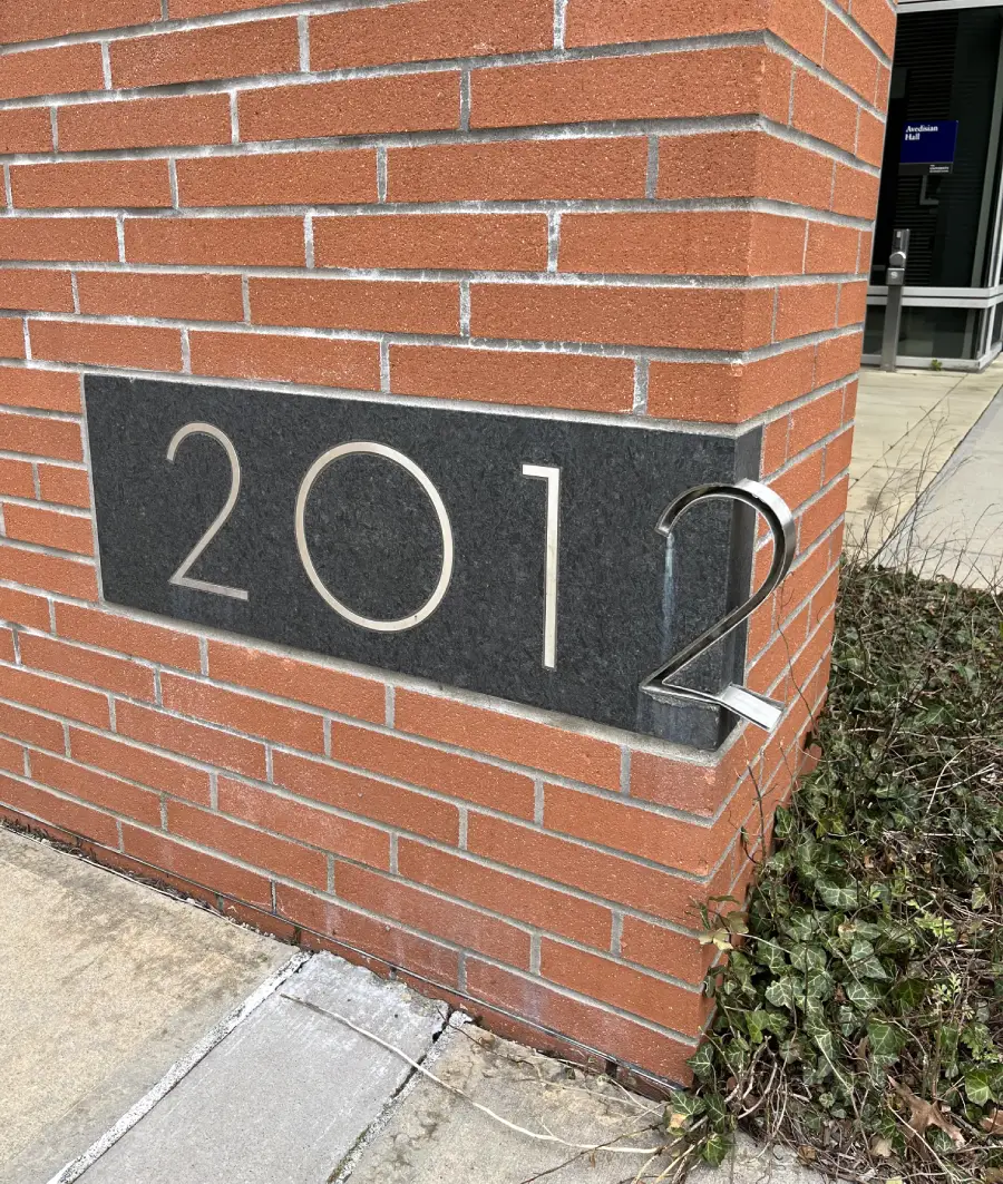 A metal plaque with the number "2012" is embedded into the corner of a red brick building, with the digit "2" extending around the edge; some ivy grows at the building's base.