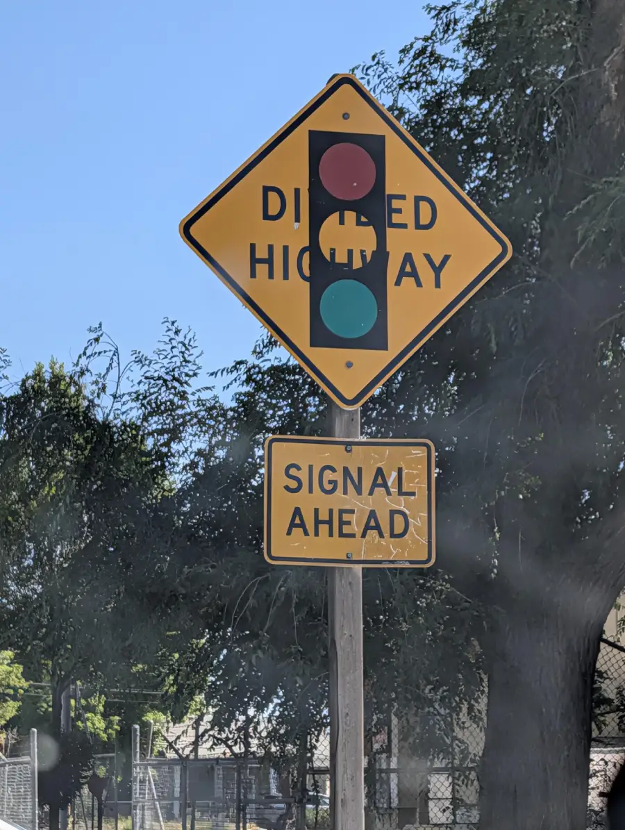 A yellow diamond-shaped road sign shows a traffic light and reads "DIVIDED HIGHWAY." Below it, a rectangular yellow sign reads "SIGNAL AHEAD." Trees and a fence are visible in the background.