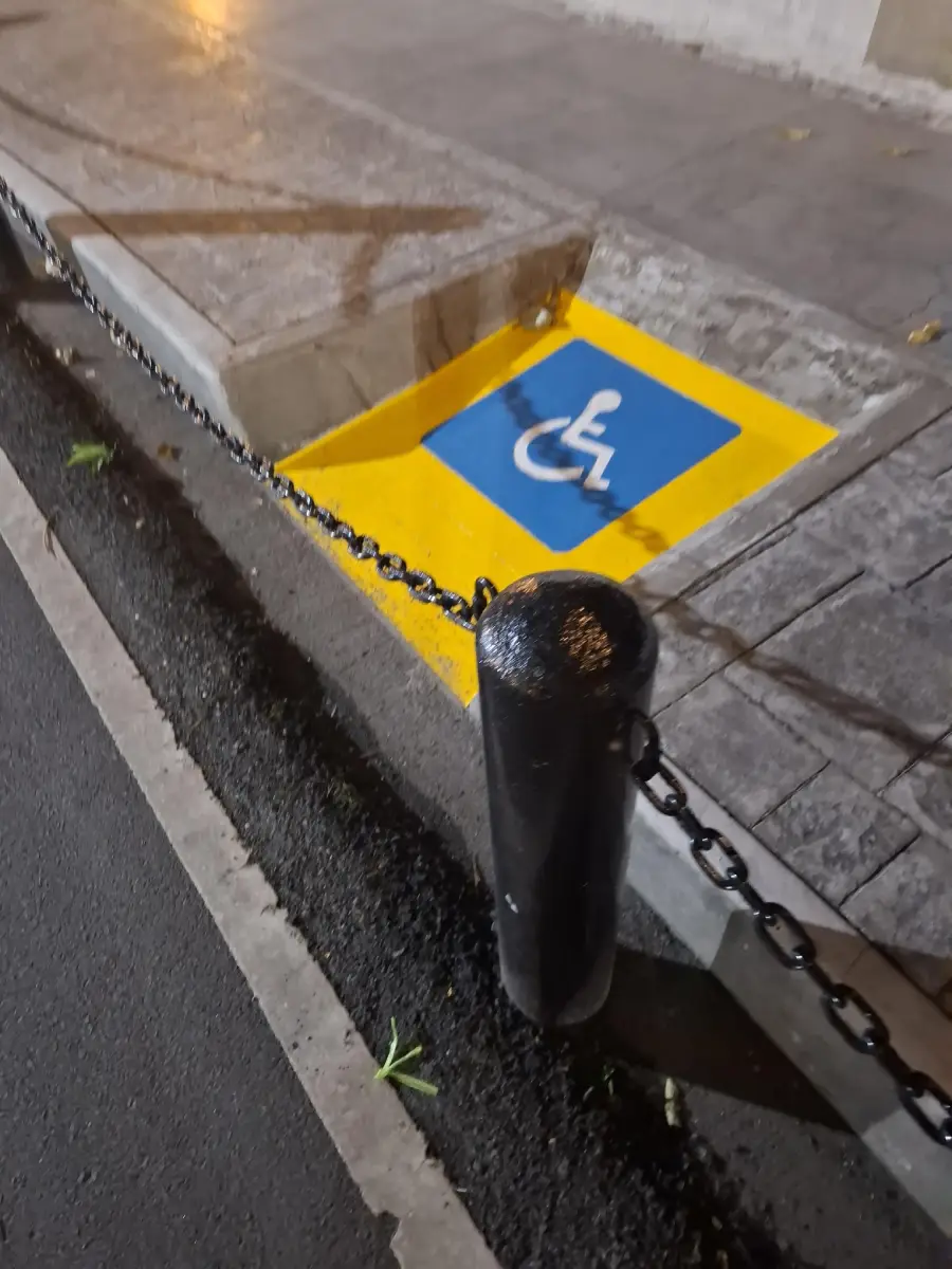A wheelchair accessible parking symbol is painted at the base of a curb with no ramp, blocked by a chain and pole, making access impossible.