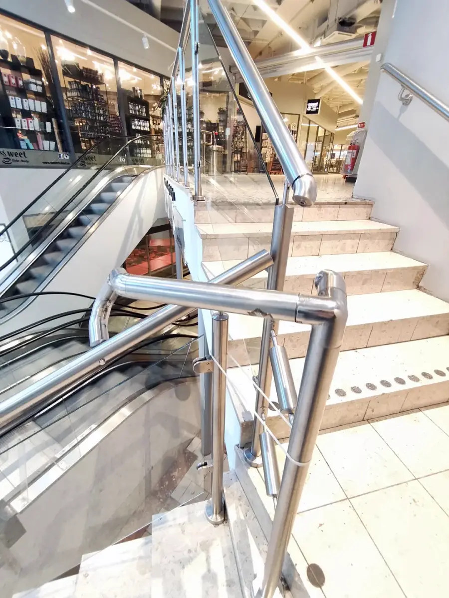 A staircase in a shopping mall is blocked by metal railings, preventing access while an escalator runs beside it. Glass storefronts are visible in the background.