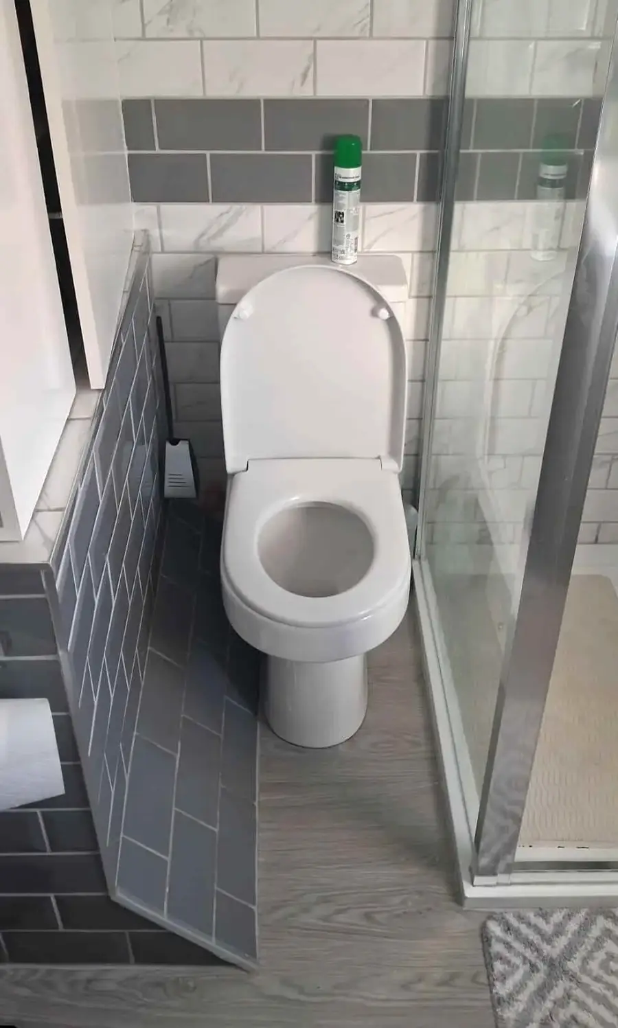 A toilet positioned in a small bathroom corner, partly blocked by a tiled wall, with a toilet brush beside it, shower enclosure to the right, and a can of air freshener on top of the cistern.