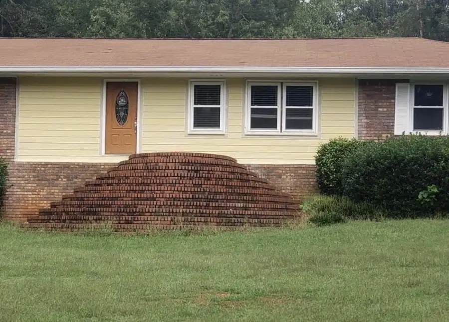 A single-story house with a tan door and steps leading up, but the brick steps form a half-circle mound with no pathway, making the entrance inaccessible. Grass and bushes surround the front yard.