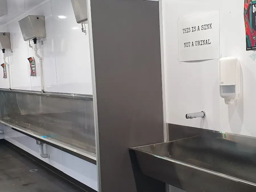 A long stainless steel sink runs along the wall, with a single sink on the right. A sign above the right sink reads, "THIS IS A SINK NOT A URINAL." The area appears to be in a public restroom or facility.