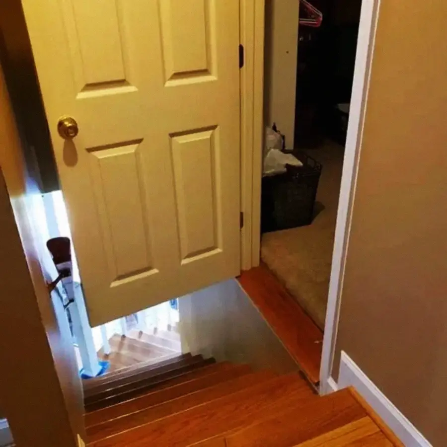 A door opens onto a staircase, but the door blocks most of the steps, creating a hazardous and awkward entryway. The stairs lead down from a small landing with tan walls and wood flooring.
