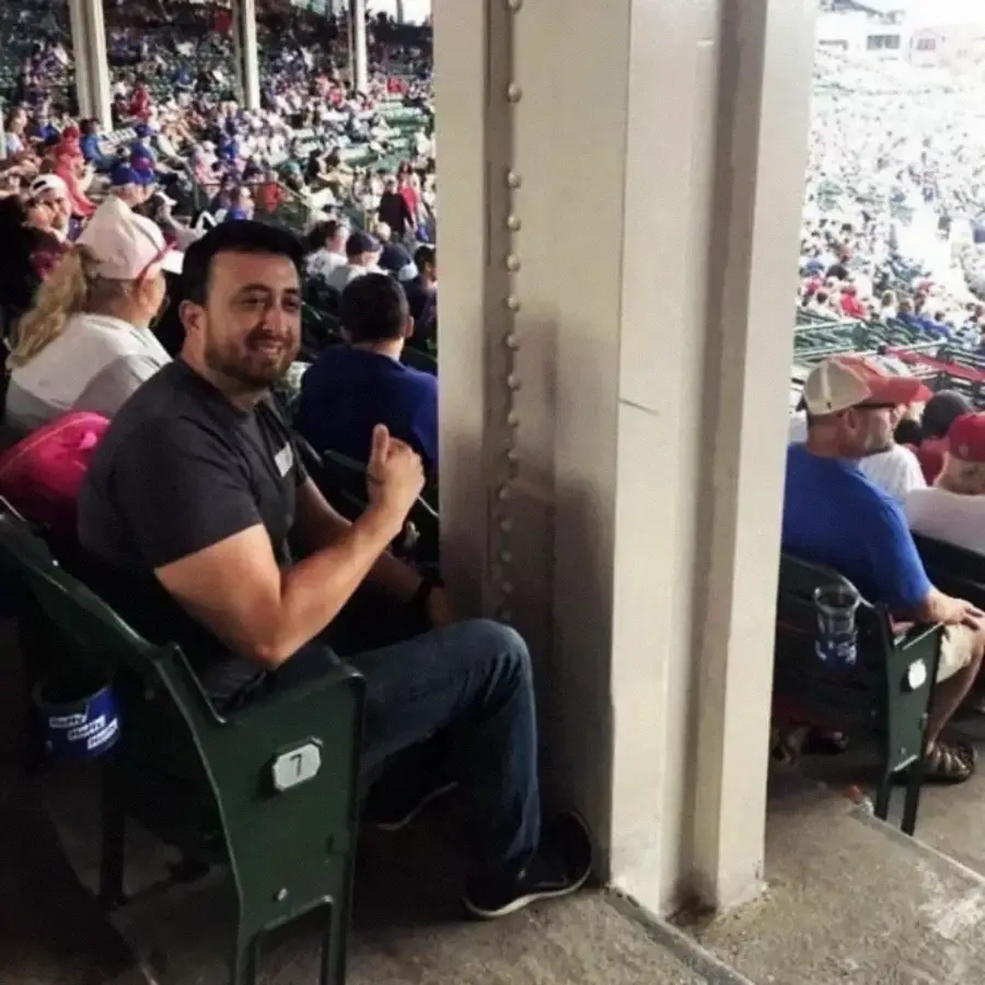 A man sits in a stadium seat directly behind a large pillar, blocking his view of the field. He smiles and gives a thumbs-up to the camera, while other spectators watch the game.