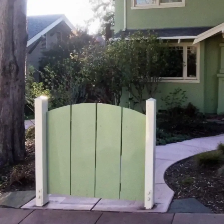 A small, light green wooden gate stands alone with no fence on either side, in front of a green house with a curved pathway and garden.