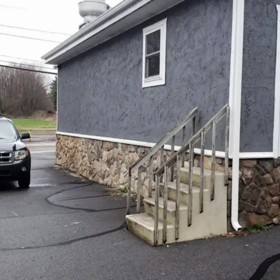 A short staircase with a metal railing leads to a blank wall with no door, next to a parking lot and a gray stone building. A parked SUV is visible on the left.
