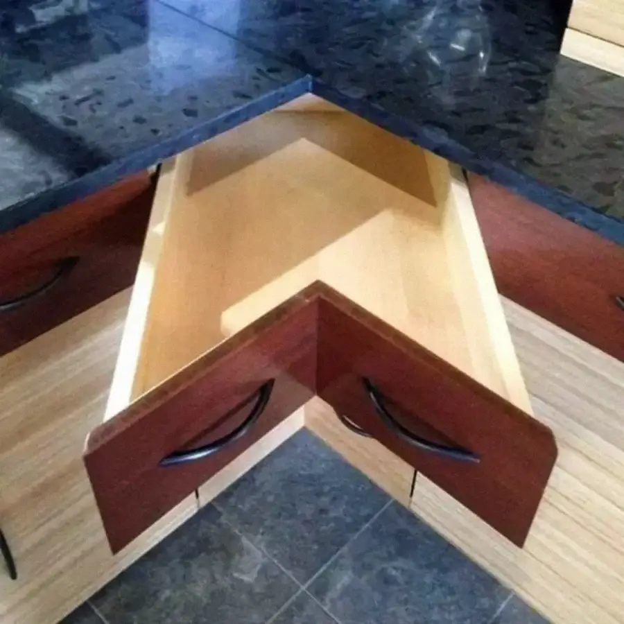 A corner kitchen drawer is open, showing an unusual triangular shape that fits into the corner of the countertop. The drawer has two handles and is surrounded by wood cabinetry and a dark countertop.