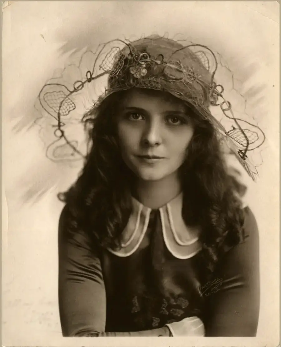 A young woman with wavy dark hair wears a vintage dress with a white collar and an elaborate, decorative hat adorned with lace and wire shapes. She looks directly at the camera with a soft expression. The photo is sepia-toned.