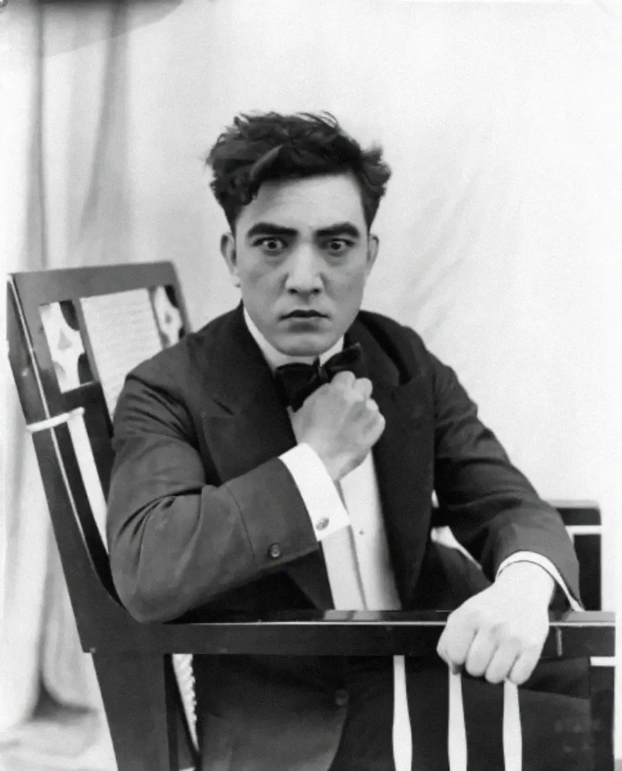 A man with tousled dark hair sits in a decorated chair, wearing a suit, bow tie, and white shirt. He looks directly at the camera with a serious expression, one hand adjusting his collar. The background is plain and light-colored.