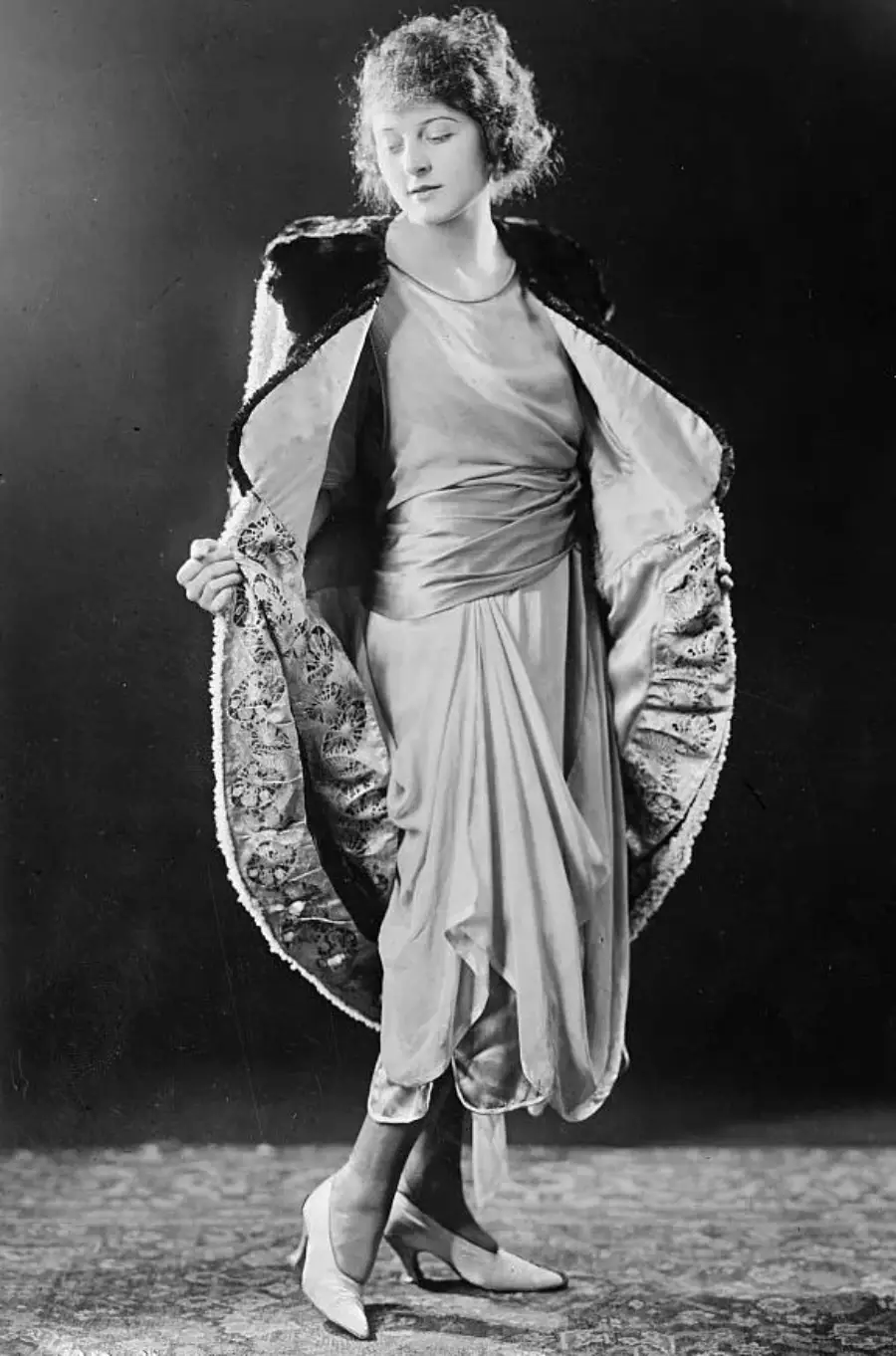 A woman in vintage 1920s attire poses with her coat open, revealing a light-colored, draped dress and patterned coat lining. She stands gracefully, looking down, against a dark backdrop.