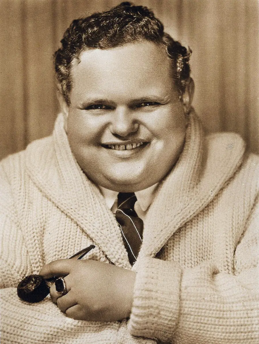 A smiling man with curly hair wears a thick knitted sweater over a shirt and tie, holding a pipe in one hand and resting his arm on a surface. The image is sepia-toned and has a vintage look.