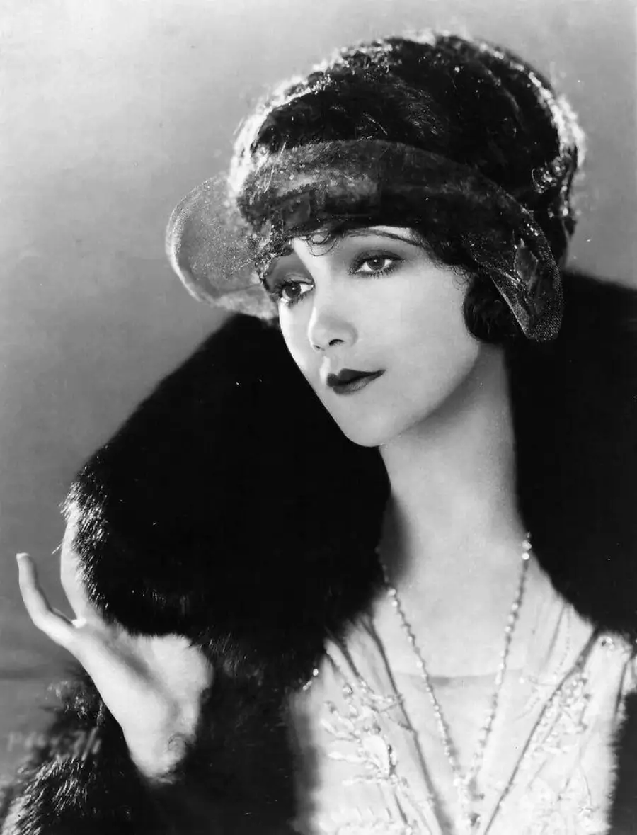 A glamorous woman from the 1920s poses elegantly, wearing a dark fur stole, a beaded dress, and a decorative headpiece with a sheer veil. Her hair is styled in soft waves and she has a serene, composed expression.