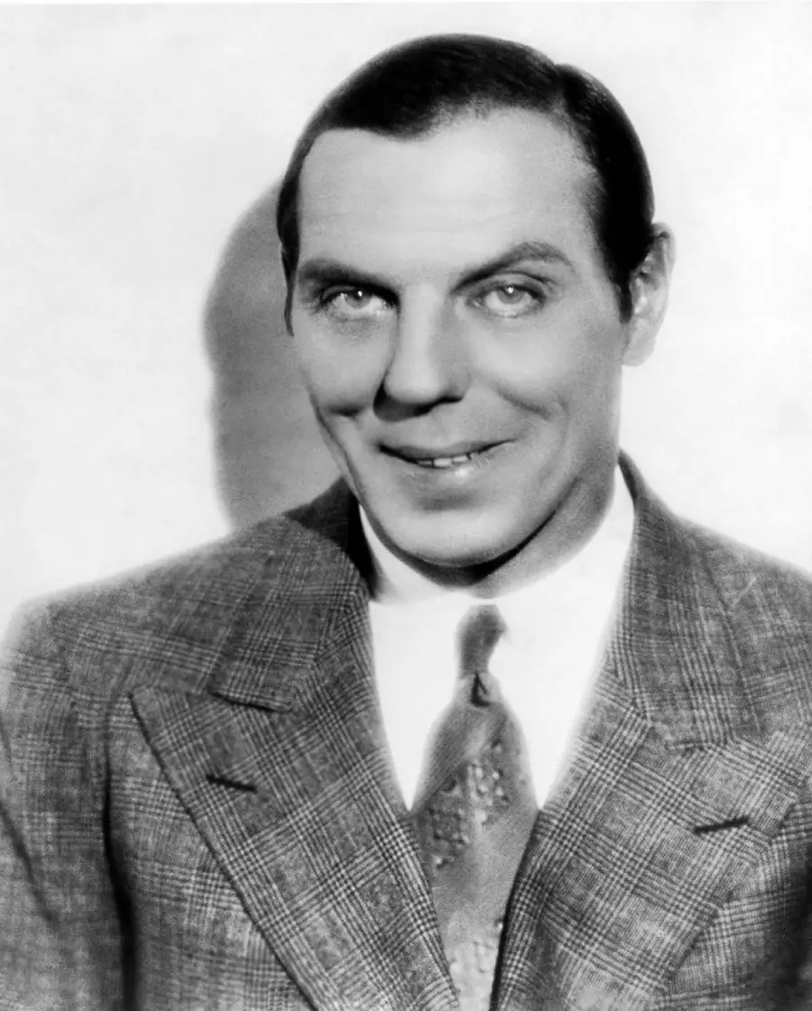 Black and white portrait of a man in a checkered suit, dress shirt, and patterned tie, smiling slightly while looking at the camera against a plain background.