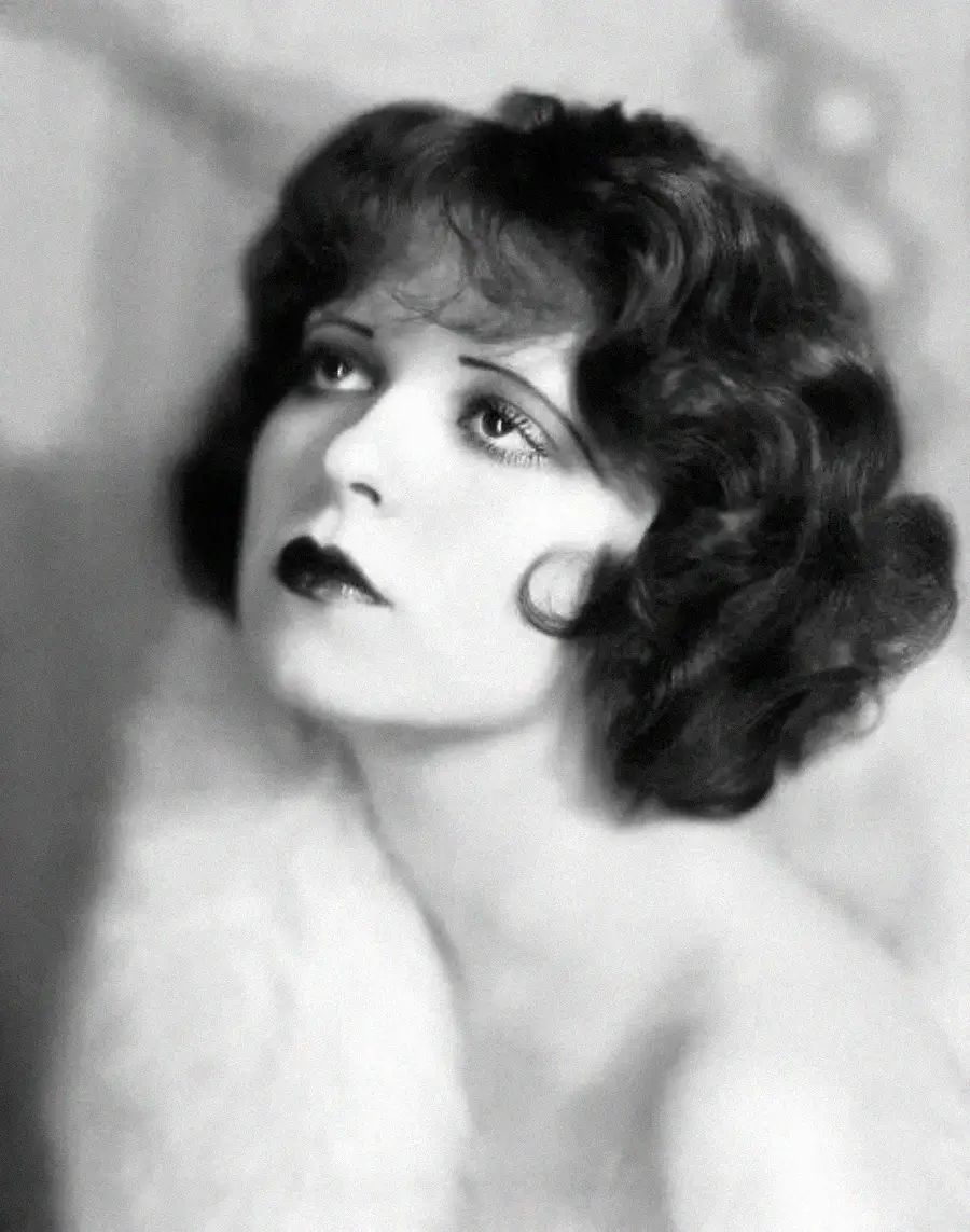 Black and white portrait of a woman with short, wavy dark hair, dark lipstick, and dramatic eye makeup, looking slightly upward. She is draped in a fuzzy, light-colored fabric.