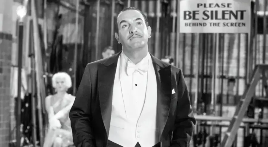 A man in a tuxedo stands with hands in pockets, looking upward. Behind him, a woman in a dress sits blurred. A sign reads "Please BE SILENT behind the screen." The scene is in black and white.