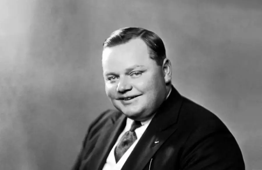 A man in a dark suit, vest, and tie poses for a formal portrait, smiling slightly. He has neatly combed hair and is set against a plain, softly lit background.