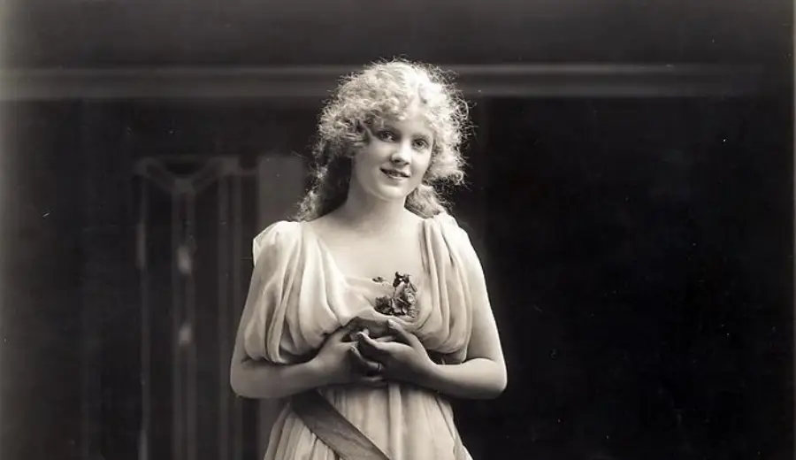 A young woman with curly hair, wearing a light, flowing dress, stands holding her hands together at her chest and looking slightly upward with a gentle expression. The background is dark and out of focus.