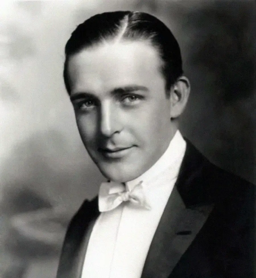 A black-and-white portrait of a man with slicked-back hair wearing a tuxedo and bow tie, looking directly at the camera with a slight smile. The background is softly blurred.