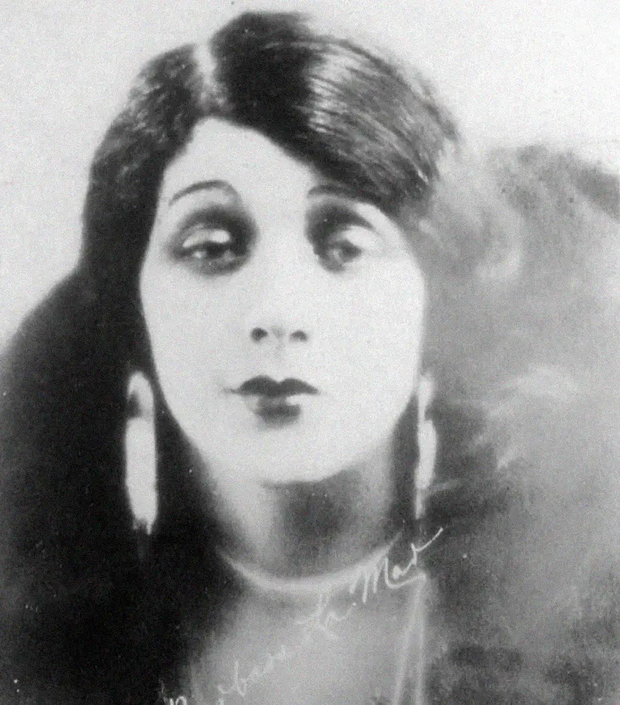 Black and white portrait of a woman with long dark hair, parted in the middle, wearing large earrings and a necklace, looking slightly to the side with a neutral expression.
