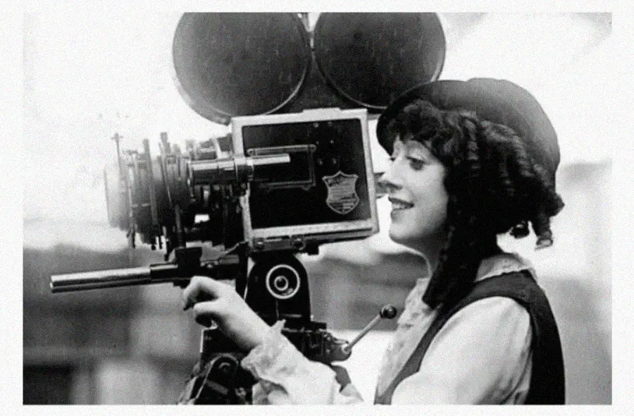 A smiling woman in early 20th-century attire operates a vintage film camera with large reels, suggesting a historical setting in the early days of filmmaking.