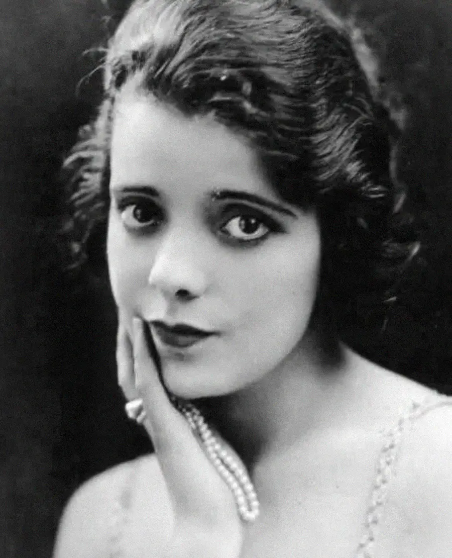 A young woman with short, wavy dark hair gazes at the camera, gently touching her face with one hand. She wears a pearl necklace and a ring, with a calm expression against a dark background.