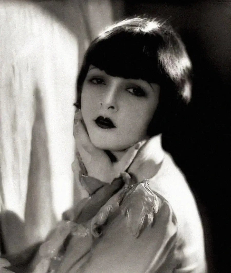 A woman with short dark bobbed hair and dark lipstick poses with her chin resting on her hand, gazing thoughtfully at the camera. The image is in black and white, giving it a classic, vintage feel.