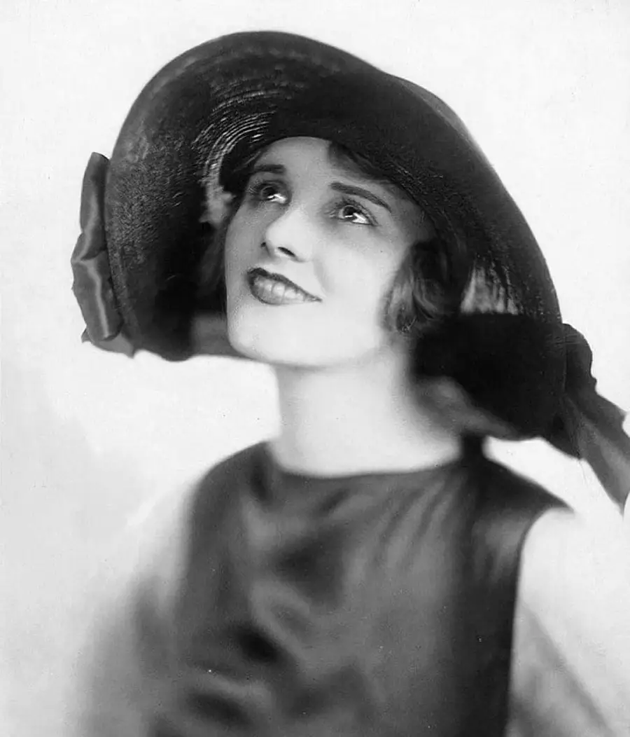 A vintage black-and-white portrait of a young woman wearing a wide-brimmed hat with a large bow. She is smiling softly and looking slightly upwards, dressed in a sleeveless top or dress.