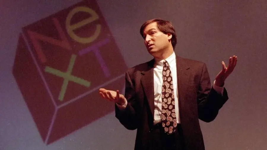 A man in a suit gestures with open hands while standing in front of a large NeXT logo projected on a screen.