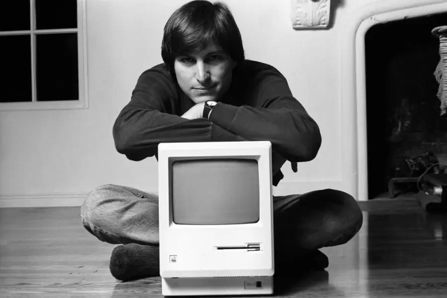 A person with medium-length hair sits cross-legged on the floor, resting their arms on top of an early model Macintosh computer, with a window and fireplace in the background. The image is in black and white.