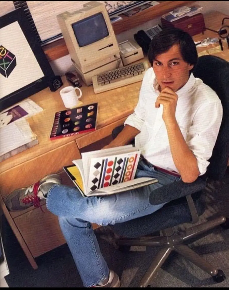 A man in jeans and a white shirt sits in an office chair, holding a book with geometric designs. He is at a desk with papers, a coffee mug, and a vintage computer, in a workspace with creative materials.