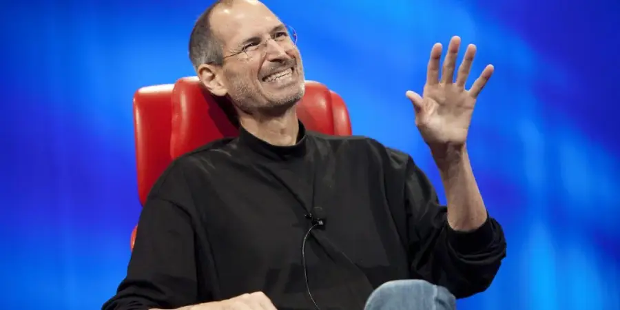 A man wearing glasses and a black long-sleeve shirt sits in a red chair, smiling and waving his hand, with a blue background behind him.