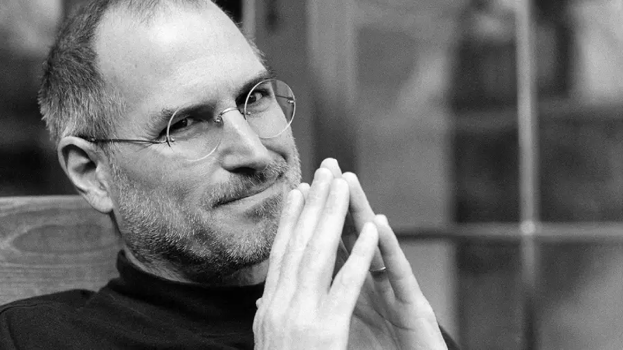 A man with glasses and a beard is sitting with his fingers pressed together near his face, wearing a dark turtleneck and smiling thoughtfully in a black and white photo.