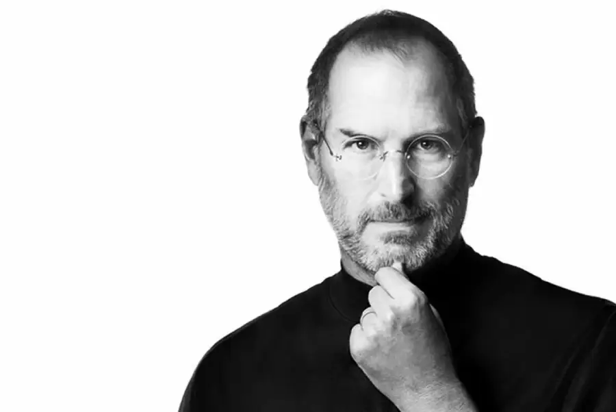 A man with glasses and a beard, wearing a black turtleneck, looks directly at the camera while resting his chin on his hand against a plain white background.